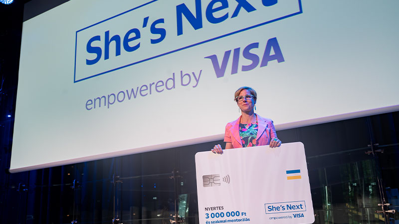 woman holding prize in front of She's Next logo on screen