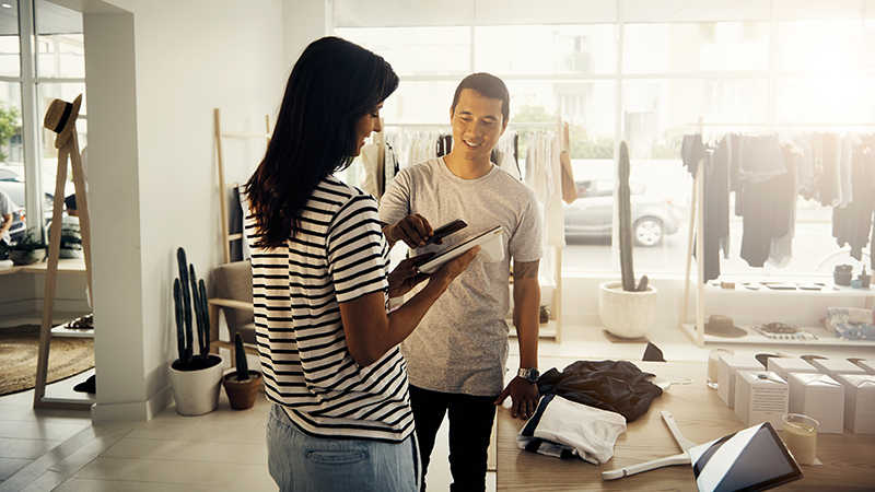 Man with mobile phone and woman with tablet completing checkout in a clothing shop.